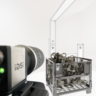 IDS uEye CP industrial camera focussed on a pallet cage with metal parts, while a modern lighting and scanning unit can be seen in the background