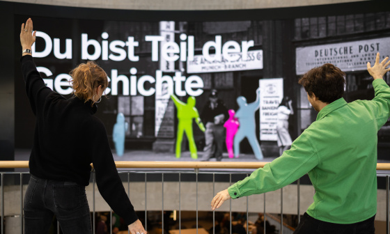 Two people are posing in front of a large interactive display wall featuring coloured silhouettes