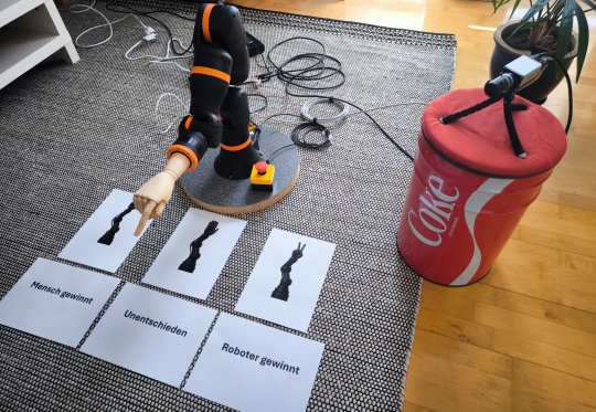 Robot arm points to sheets of paper in front of it, IDS NXT camera stands next to it