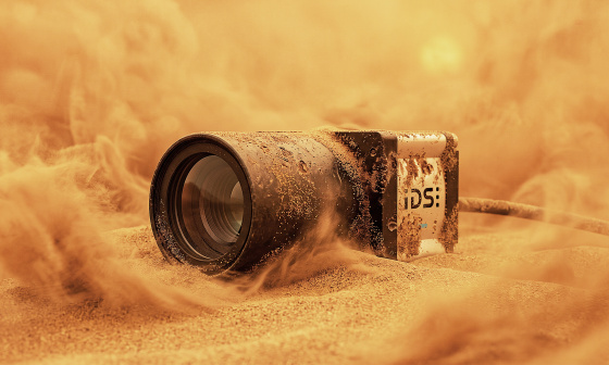 Industrial camera partly buried in sand, surrounded by swirling sand.