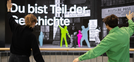 Two people are posing in front of a large interactive display wall featuring coloured silhouettes